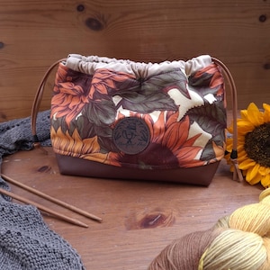 Könnte beinhalten: Ein Projektbeutel mit Kordelzug, verziert mit einem Sonnenblumenmuster in Orange, Gelb und Grün, sowie einem Boden aus braunem Kunstleder. Der Beutel hat einen beigen Stoffabschluss, braune Kordeln und ein rundes, dunkelbraunes Logo mit Hexenhut.