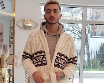 May include: A white knitted cardigan with a brown and white geometric pattern. The cardigan has a hood and a tie belt. The person is wearing dark blue pants and white sneakers.