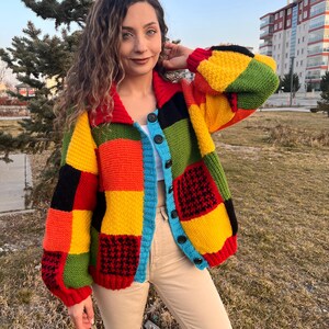 Könnte beinhalten: Ein bunter Patchwork-Cardigan mit Knopfleiste. Der Cardigan ist aus dickem Strickstoff gefertigt und in verschiedenen Farben gehalten, darunter Rot, Gelb, Grün, Blau und Schwarz.