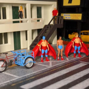 May include: Three miniature figures wearing red capes and blue costumes stand in front of a motel. One figure is wearing a blue mask. A small tricycle is in the foreground.