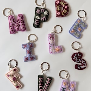 May include: A collection of ten felt letter keychains in various colours including lavender, dark green, burgundy, and cream. Each letter features intricate floral and vine embroidery in contrasting thread colours and is attached to a silver metal ring.
