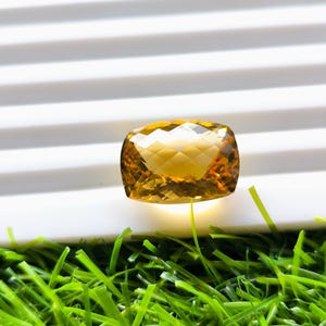 Peut inclure: Une pierre précieuse jaune doré, taillée en coussin rectangulaire. La gemme est posée sur un fond blanc strié et de l'herbe artificielle verte brillante.