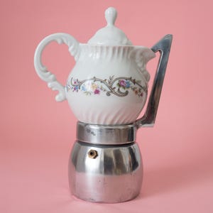 May include: A white porcelain coffee pot with floral detailing sits atop a silver stovetop espresso maker. The pot has a curved handle and a lid with a small knob. The background is a solid pink.