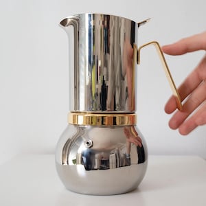 Puede incluir: Una cafetera italiana de acero inoxidable con asa y detalles dorados. La cafetera tiene una base bulbosa y una parte superior delgada con un pico vertedor. El metal pulido refleja el entorno.