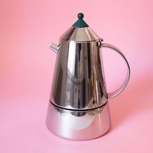 Puede incluir: Una cafetera de acero inoxidable pulido con cuerpo cónico, asa curva y un pequeño pico vertedor. La tapa presenta un pomo redondeado de color verde oscuro en la parte superior. La superficie metálica es reflectante y brillante.