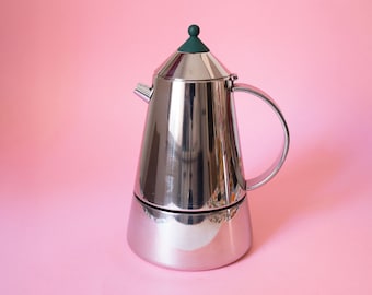 Könnte beinhalten: Ein polierter Espressokocher aus Edelstahl mit konischem Körper, einem geschwungenen Griff und einem kleinen Ausguss. Der Deckel ist mit einem dunkelgrünen, abgerundeten Knopf versehen. Die Metalloberfläche ist glänzend und reflektierend.