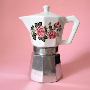 Peut inclure: Une cafetière italienne en céramique blanche avec un motif de roses et une base argentée. La poignée et le couvercle sont blancs. Les roses sont roses et rouges avec des feuilles vertes.
