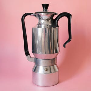 Pode incluir: Uma cafeteira de alumínio prateado para fogão com corpo cilíndrico, alça de plástico preto e um botão preto correspondente na tampa. A cafeteira possui uma base larga, uma seção central mais estreita e um bico para servir o líquido.