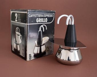 May include: A silver and black espresso maker with a chrome finish. The maker is labeled "Caffettiera Espresso Grillo" on the box. The box also states "2 Tazze Cups Tazas Tassen Tassen".