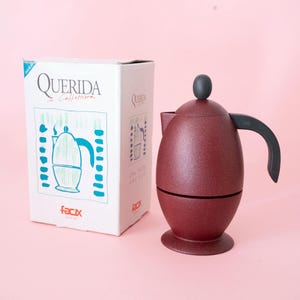 Pode incluir: Uma cafeteira de cor vermelho-escura em forma de ovo, com pega e botão da tampa pretos, ao lado da sua embalagem de cartão branca. A caixa apresenta o texto "Querida Caffettiera" e "facix" com uma ilustração azul e verde de uma cafeteira.
