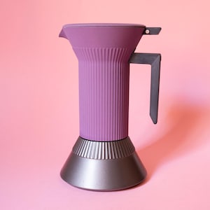 Pode incluir: Uma cafeteira espresso roxa com uma textura canelada vertical no corpo superior. A base é de formato cônico cinza-escuro liso com uma transição canelada correspondente. Possui uma alça angular cinza-escura e uma tampa articulada no topo.