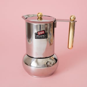 May include: A stainless steel stovetop espresso maker with a polished silver finish. It features a gold-toned cylindrical handle and a matching gold knob on the lid. A black label on the front displays the white and red text VEV VIGANO.