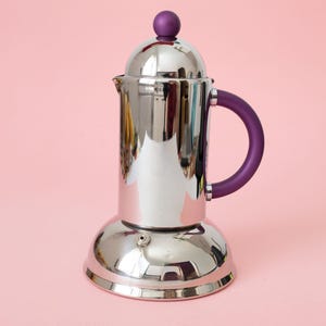 May include: A polished stainless steel stovetop espresso maker with a wide, rounded base and a cylindrical body. It features a matte purple handle and a matching purple spherical knob on the domed lid. A small metal pressure valve is visible on the base.