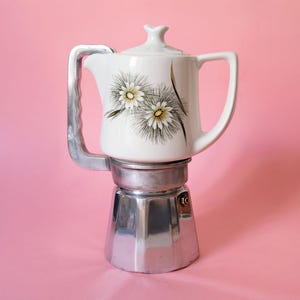 以下が含まれることがあります： 白い陶器製の上部チャンバーを備えた直火式エスプレッソメーカー。上部には白い花びらと茶色の棘のような模様が描かれています。ベースとサイドハンドルは磨かれた銀色の金属製で、小さな圧力バルブが付いています。