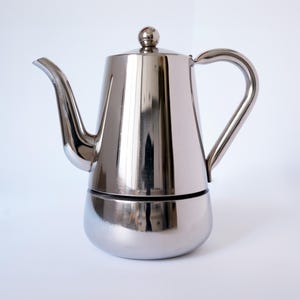 May include: A shiny, stainless steel stovetop espresso maker. The coffee pot has a rounded base, a cylindrical upper chamber, a curved spout, and a loop handle. The lid is topped with a small, round knob.