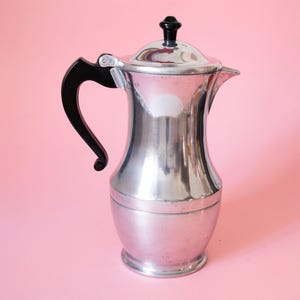 Puede incluir: Una cafetera de metal color plata con acabado pulido, que presenta un asa negra curvada, una tapa con bisagra con un pequeño pomo negro y un pico corto. El cuerpo tiene una base redondeada que se estrecha hacia el cuello antes de ensancharse.