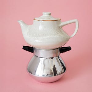 May include: A white ceramic teapot with a speckled finish and a matching lid sits atop a metallic silver stovetop espresso maker. The espresso maker features a rounded base, a central silver band, and two black plastic handles extending outward.