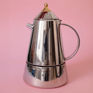 Puede incluir: Una cafetera espresso de acero inoxidable pulido con cuerpo cónico, asa curva y un pequeño pico vertedor. La tapa presenta una parte superior puntiaguda con un pequeño pomo esférico de color dorado. La superficie es metálica y muy reflectante.