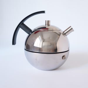 Könnte beinhalten: Eine kugelförmige Teekanne aus Edelstahl mit schwarzem Griff und Ausguss. Die Teekanne hat einen kleinen Deckel und eine polierte, reflektierende Oberfläche. Das Design ist modern und minimalistisch. Durchmesser ca. 20 cm.