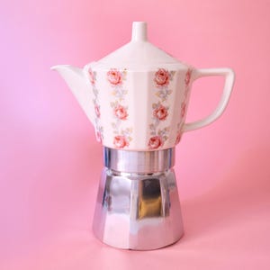 Könnte beinhalten: Ein Espressokocher mit einem weißen Keramikoberteil, das eine vertikal gerippte Textur und ein florales Muster aus rosa Rosen und grünen Blättern aufweist. Der untere Teil besteht aus poliertem silberfarbenem Metall mit einem passenden Keramikdeckel.