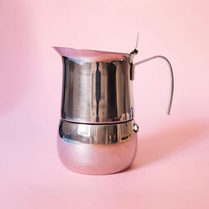 Puede incluir: Una cafetera espresso de acero inoxidable pulido con base redondeada, cámara superior cilíndrica, asa curva y un pequeño pico vertedor. La superficie metálica refleja el entorno, mostrando un acabado claro y brillante.