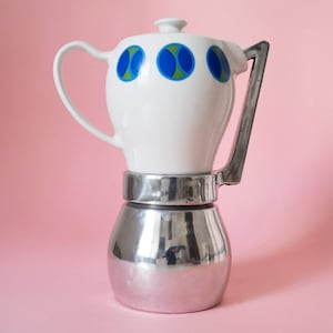 Może przedstawiać: Biały ceramiczny ekspres do kawy na kuchenkę z srebrną podstawą i uchwytem. Górna część ma trzy niebieskie i zielone okrągłe wzory. Uchwyt jest w kolorze ciemnoszarym. Ekspres do kawy stoi na różowym tle.