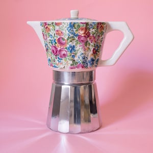May include: A stovetop espresso maker featuring a metallic silver base with a faceted design and a white ceramic upper chamber decorated with a colourful floral pattern of pink roses and blue flowers, complete with a matching white lid and handle.