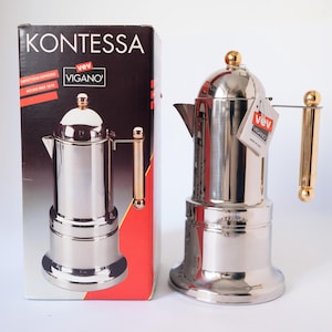 Op de afbeelding: Een roestvrijstalen espressomaker met een goudkleurige handgreep en accenten, naast de originele doos. De doos toont de merknaam "KONTESSA" en een afbeelding van de espressomaker. De espressomaker heeft een klassiek ontwerp.