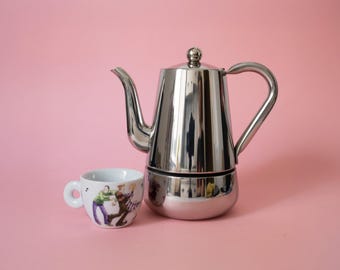Könnte beinhalten: Ein polierter Espressokocher aus Edelstahl mit hohem, konischem Körper, geschwungenem Ausguss und passendem Griff, neben einer weißen Espressotasse aus Keramik, die mit einer farbenfrohen grafischen Illustration von zwei tanzenden Personen verziert ist.