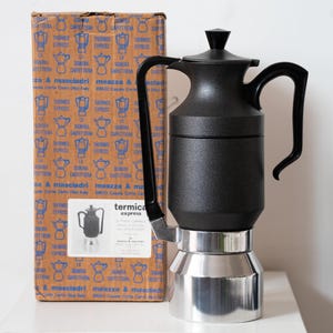Könnte beinhalten: Ein schwarzer und silberner Termica Express Espressokocher mit schwarzem Griff und Deckel. Der Espressokocher steht neben seinem Originalkarton mit blauem und braunem Design.