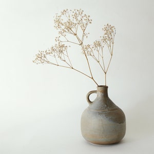 May include: A gray ceramic vase with a textured surface and a handle, holding dried white flowers.