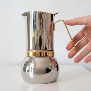 May include: A stainless steel stovetop espresso maker with a gold-colored handle and band. The coffee pot has a rounded base and a spout. The handle is held by a hand.