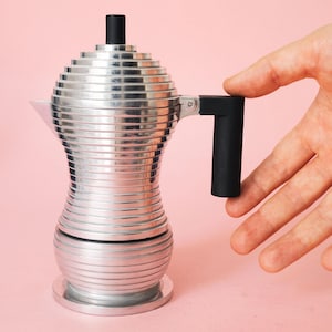 Puede incluir: Una cafetera moka de aluminio plateado con una textura de nervaduras horizontales en todo el cuerpo, un mango cilíndrico negro y un pomo negro a juego en la tapa. La cafetera tiene una base redondeada y una sección superior cónica.