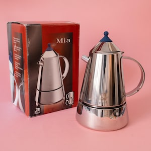 May include: A polished stainless steel stovetop espresso maker with a conical body, a curved handle, and a small blue knob on the lid. Next to it stands its original rectangular cardboard box, which features the product name Mia and a graphic image.