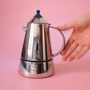 以下が含まれることがあります： 円錐形のボディ、湾曲したハンドル、蓋に小さな青いノブが付いた、磨き上げられたステンレス製の直火式エスプレッソメーカー。コーヒーポットは反射する金属仕上げと特徴的な注ぎ口を備えており、洗練されたモダンなデザインが際立っています。
