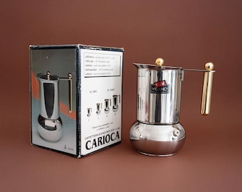 May include: A stainless steel stovetop espresso maker with a gold-colored handle and accents, next to its original box. The box features an illustration of the coffee maker and text in Italian. The coffee maker is designed to brew 4 cups.