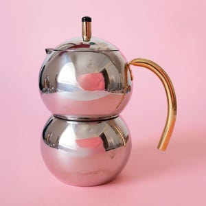 May include: A polished silver-tone metal teapot with a spherical body, a curved gold-tone handle, and a small lid featuring a black knob. The reflective surface shows a pink tint, and the design consists of two stacked rounded sections.