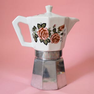 Puede incluir: Una cafetera italiana blanca y plateada con un diseño floral de rosas. La parte superior es blanca con un asa y un pico, mientras que la base es plateada. Las rosas son en tonos rosa y naranja con hojas verdes.