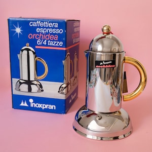 Pode incluir: Uma cafeteira espresso de aço inoxidável polido com uma alça dourada e um pequeno botão dourado na tampa. A cafeteira possui um rótulo preto com o texto Inoxpran orchidea. É exibida ao lado de sua caixa de papelão azul com texto branco.