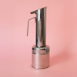 May include: A polished stainless steel oil dispenser featuring a cylindrical base, a tall vertical body, and a side handle. The dispenser includes a top spout mechanism designed for controlled pouring, with a reflective metallic finish throughout.
