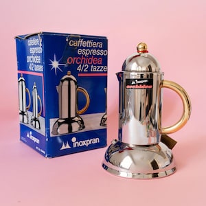 Puede incluir: Una cafetera espresso de acero inoxidable pulido con un asa de tono dorado y un pequeño pomo dorado en la tapa. La cafetera presenta una base ancha y redondeada y una etiqueta negra con el texto "Inoxpran orchidea". Se muestra junto a su caja azul.