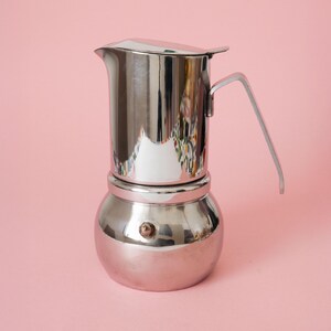 May include: A polished stainless steel stovetop espresso maker with a cylindrical upper chamber, a rounded lower water reservoir, a hinged lid, and a slender angular handle. A small metal pressure release valve is visible on the lower base.