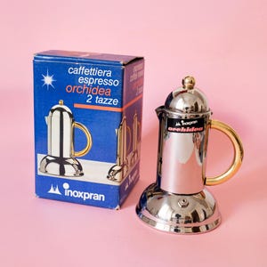 Pode incluir: Uma cafeteira espresso Inoxpran Orchidea de aço inoxidável polido com alça dourada e a sua caixa de cartão azul. A caixa apresenta o texto caffettiera espresso orchidea 2 tazze e o logótipo Inoxpran na parte frontal e lateral.