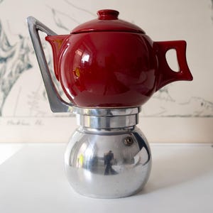 Puede incluir: Una tetera de cerámica roja con asa y tapa plateadas se asienta sobre una cafetera plateada. La cafetera tiene una base redondeada. El fondo presenta una obra de arte enmarcada.