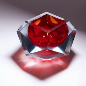 Puede incluir: Un cuenco de cristal facetado con una carcasa exterior transparente y angular y un interior rojo intenso, liso y redondeado. El diseño geométrico crea bordes afilados y superficies planas que refractan la luz, resaltando el contraste entre el exterior transparente y el centro rojo vibrante.