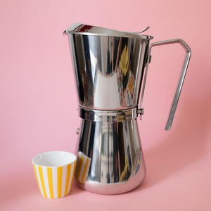 Könnte beinhalten: Ein Espressokocher aus poliertem Edelstahl mit hohem, konischem Körper und seitlichem Griff, neben einer kleinen weißen Keramiktasse mit vertikalen gelben Streifen auf der Außenseite.