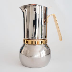 May include: A stainless steel espresso maker with a gold-coloured handle and band. The coffee pot has a rounded base and a tall, cylindrical top. The polished metal reflects the surrounding environment, creating a shiny appearance.