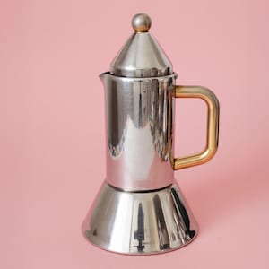 Op de afbeelding: Een gepolijste zilverkleurige metalen espressopot voor op het fornuis met een conisch deksel bekroond door een kleine bolvormige knop. Het cilindrische lichaam heeft een brede, uitlopende voet en een gladde, goudkleurige metalen handgreep.