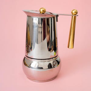 Pode incluir: Uma cafeteira de aço inoxidável polido com base arredondada e corpo superior cônico. A tampa possui um pequeno botão esférico dourado e a alça lateral é um cilindro longo e fino de cor dourada com uma esfera correspondente.