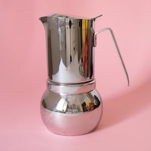 May include: A shiny, stainless steel stovetop espresso maker with a curved handle. The coffee pot has a bulbous base and a pouring spout. The background is a solid pink.